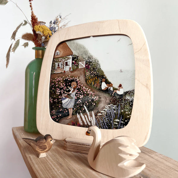 Wooden picture frame with a painted scene of a garden and house, placed on a wooden surface with decorative items.